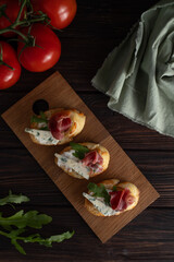 On the kitchen table is a board with fresh bruschetta with ham and cheese. Along with tomatoes and greens