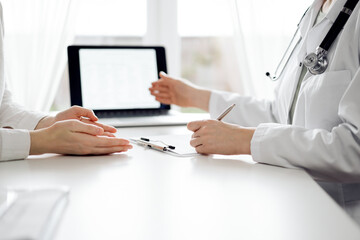Doctor and patient sitting and discussing something near each other at the white desk in clinic. Female physician is pointing into laptop screen. Medicine concept