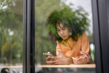 Portrait photo from outside the window glass of Young African American woman using mobile phone...
