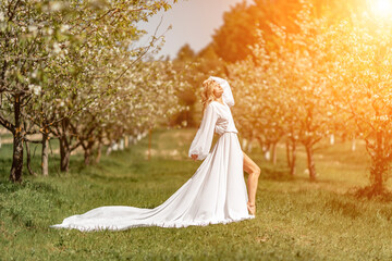 Fototapeta na wymiar Blond blooming garden. A woman in a white dress walks through a blossoming cherry orchard. Long dress flies to the sides,