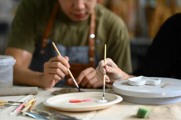Concentrated man painting clay plate with a paintbrush at desk in ceramic workshop