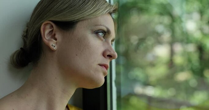 Young woman alone at home thinks about loneliness or grief and sits window and looks out into street with lonely expression. Loneliness depression and stress