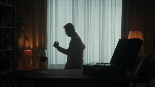 Vintage record player with a rotating vinyl record and a glass of red wine on the table close up. A man dances alone, bowing. The dark silhouette of a dancing man in the room. Slow motion.