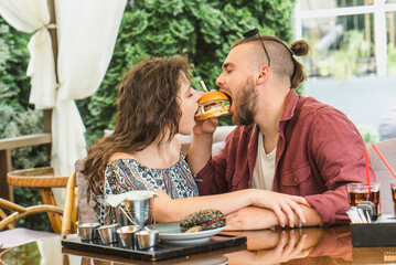 young couple sits in cafe and eats burgers with cola. Man and woman have a great time and relax enjoy food in cafe. Trendy wearing boy and girl have lunch. Teenager date