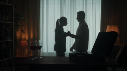 Vintage record player with a rotating vinyl record and glasses with red wine on the table close up. A couple is dancing a slow dance while enjoying a pleasant evening together. Silhouette. Slow motion