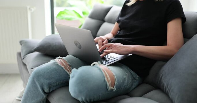 Woman uses laptop, types on keyboard and writes email sitting on couch. Person types message on social network or searches for information on Internet