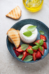 Blue bowl with ricotta cheese, sliced strawberries and fresh green basil, vertical shot on a beige stone background, elevated view