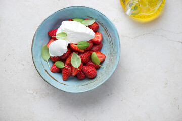 Fresh strawberry with mascarpone and green basil in a turquoise bowl, top view on a light-beige stone background, horizontal shot with space