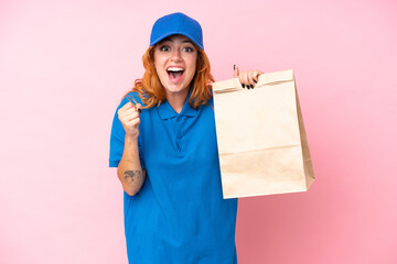 Young caucasian woman taking a bag of takeaway food isolated on pink background celebrating a victory in winner position