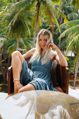 Beautiful caucasian young woman with blond hair in grey sundress sits in wooden armchair among tropical green palm trees smiling at camera. Happy girl enjoying her summer vacation. Summer trip concept