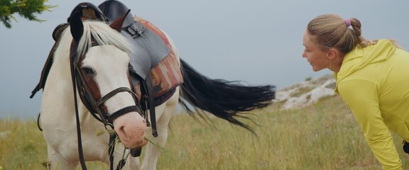 Beautiful moment human and animal. The woman traveler relax and looks at beautiful white horse. Love for pets.Mountain hill in steppe. Relaxing and interaction with nature and domestic horse outdoors