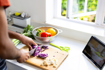 Midsection of biracial man chopping vegetables and using tablet in sunny kitchen