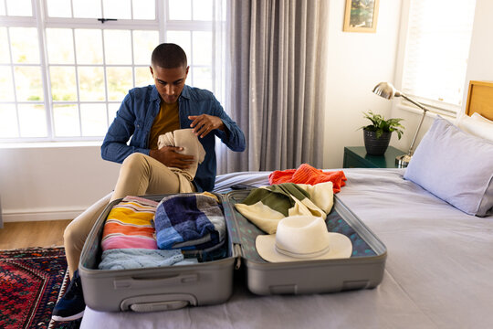 Biracial man sitting on bed packing suitcase in sunny bedroom