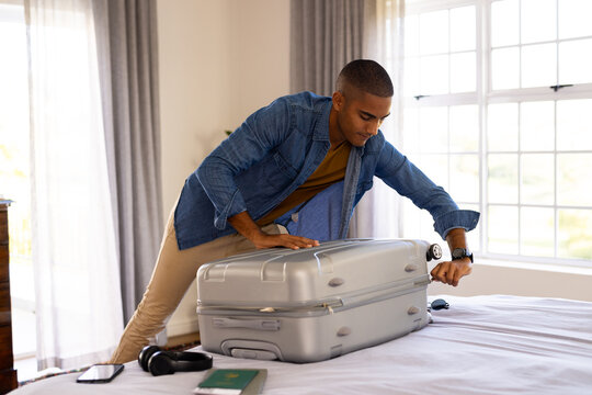Biracial man closing packed suitcase on bed in sunny bedroom