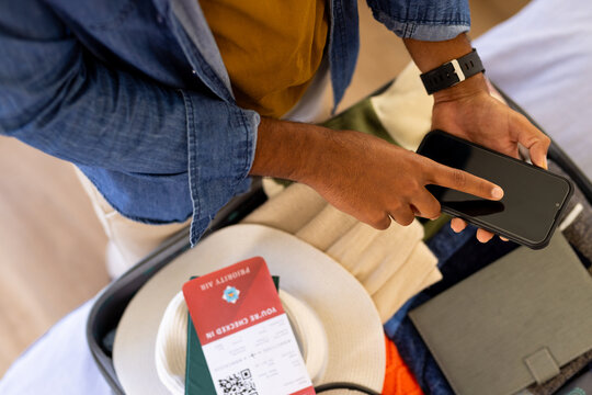 Midsection of biracial man with packed suitcase checking flight boarding pass using smartphone