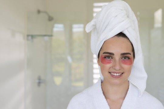 Portrait of happy caucasian woman with towel on head applying eye patches in bathroom, copy space