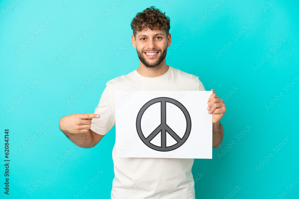 Canvas Prints Young handsome caucasian man isolated on blue background holding a placard with peace symbol and  pointing it