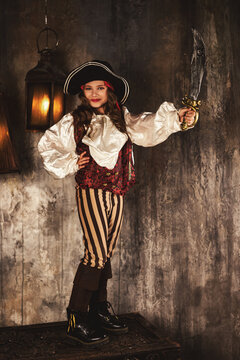 Full length of pirate showing with saber in underground room or hold of pirates ship, playful looking at camera. Cute child girl actress in corsair image. Theatre performance concept. Copy text space