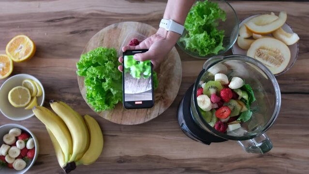 Woman  take photo on smartphone of fruits and berries on table. Healthy smoothie ingredients on table and blender bowl, top view. Various fruits , vegetables and berries. 