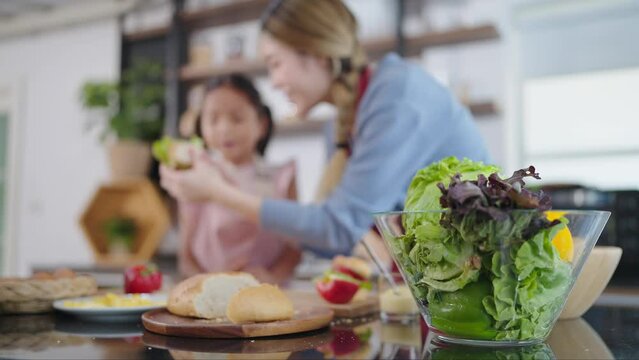 Close up vegetables salad in glessed bowl. Focus shot of vegetables salad in glessed bowl and mother and daughter helping together making sandwiches on blurred background. Healthy food concept