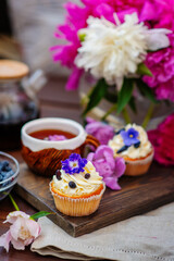 Cupcake with cream decorated with violet flowers lying next to a mug of tea among the flowers of peonies and scattered honeysuckle berries