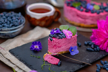 A piece of berry cheesecake lying next to a mug of tea, violet flowers and on a background of...