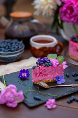 A piece of berry cheesecake lying next to a mug of tea, violet flowers and on a background of peonies and a whole cake