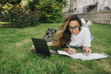 Young woman student studying using her books, laptop and notebook, sitting on grass.