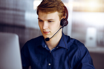 Young blond businessman using headset and computer in a darkened office, glare of light on the background. Startup business means working hard