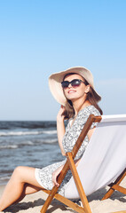 Happy brunette woman wearing sunglasses and hat relaxing on a wooden deck chair at the ocean beach - 620860771