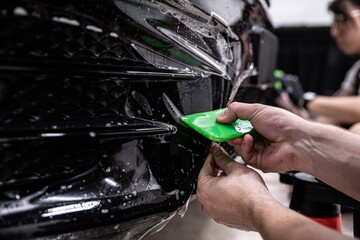 Car detailing studio employee, a wrapper, is protecting the bumper of a black car with a colorless...