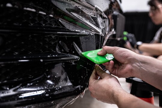 Car detailing studio employee, a wrapper, is protecting the bumper of a black car with a colorless protective PPF film. The PPF film protects the paint from scratches and damage.