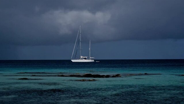 Sailing yacht at anchor in a tropical paradise with storm clouds brewing in the distance and a shallow reef in front