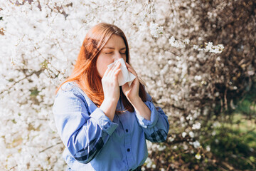 Sneezing young redhead woman with nose wiper among blooming trees in park. Portrait of sick women sneezes in white tissue, suffers from rhinitis and running nose. Symptoms of cold or allergy. - obrazy, fototapety, plakaty