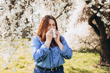 Sneezing young redhead woman with nose wiper among blooming trees in park. Portrait of sick women sneezes in white tissue, suffers from rhinitis and running nose. Symptoms of cold or allergy.