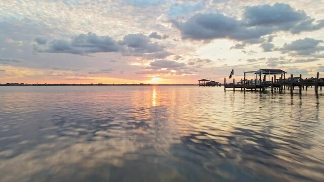 A low flying drone shot near the water. A fish can be seen hopping out of the water. Drone keeps flying by near fishing piers and docks and heads out into the ocean at sunset time.