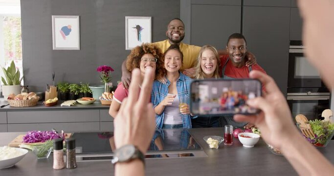 Biracial man using smartphone and taking picture of diverse friends in kitchen, slow motion