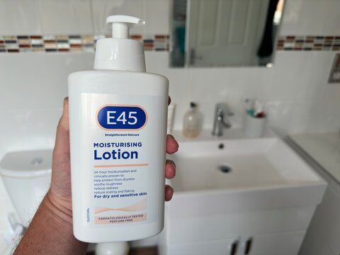 Essex, UK - July 5, 2023: E45 moisturising lotion in a plastic pump bottle being held up against a defocused  domestic bathroom. 