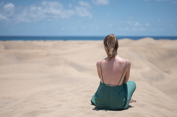 Blonde young girl in green dress sitting on sand of beach dunes