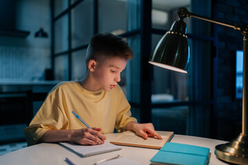 Focused redhead pupil student boy studying at home writing in exercise book doing homework, learning sitting at table under light of lamp at night. Clever schoolkid reading textbook sitting at desk.