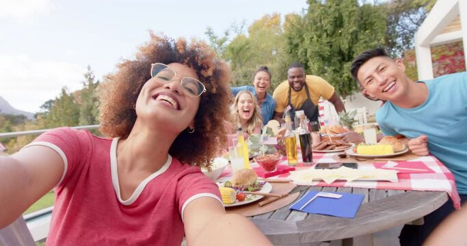 Happy diverse group of friends taking selfie at dinner table in garden, slow motion