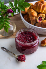Glass jar with homemade edible rose jam and crescents with rose jam on marble-wooden board.