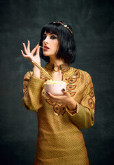 Portrait of beautiful young woman in image of egyptian queen, Cleopatra eating instant noodles against dark vintage background. Concept of antique culture, history, comparison of eras, art, beauty, ad