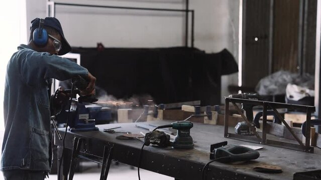 Slow motion track right of young black male working angle grinder power tool with sparks over workshop table