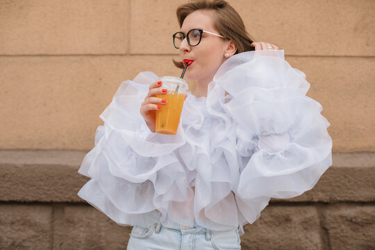 Glamorous woman in a ruffled blouse standing outdoors drinking a fruit drink