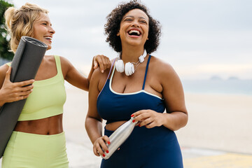 Summer fitness fun: Two women laughing and holding a yoga mat and a water bottle on the beach