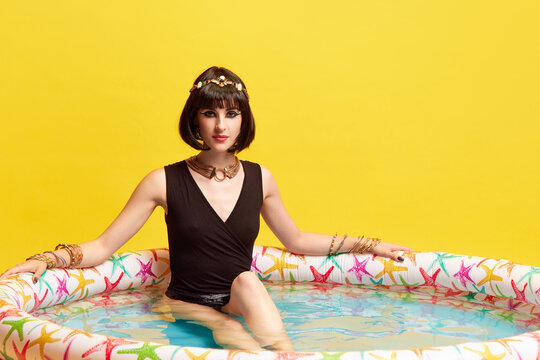 Attractive, beautiful young woman with expressive makeup, in image of Cleopatra sitting in swimming pool against yellow studio background. Concept of antique culture, history, comparison of eras, art