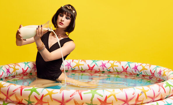 Young artistic woman in image of queen, Cleopatra in swimming pool and pouring milk inside against yellow studio background. Concept of antique culture, history, comparison of eras, art, beauty, ad