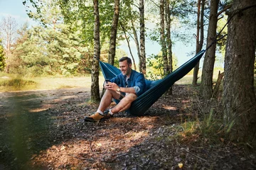 Tuinposter Man in forest sitting on hammock using mobile phone © baranq