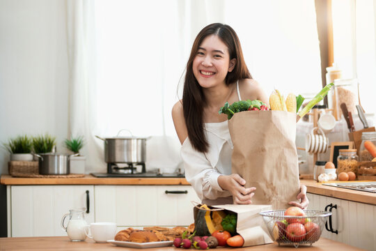 Happy Young Asian Woman In The Kitchen And Grocery Shopping Bag With Vegetables